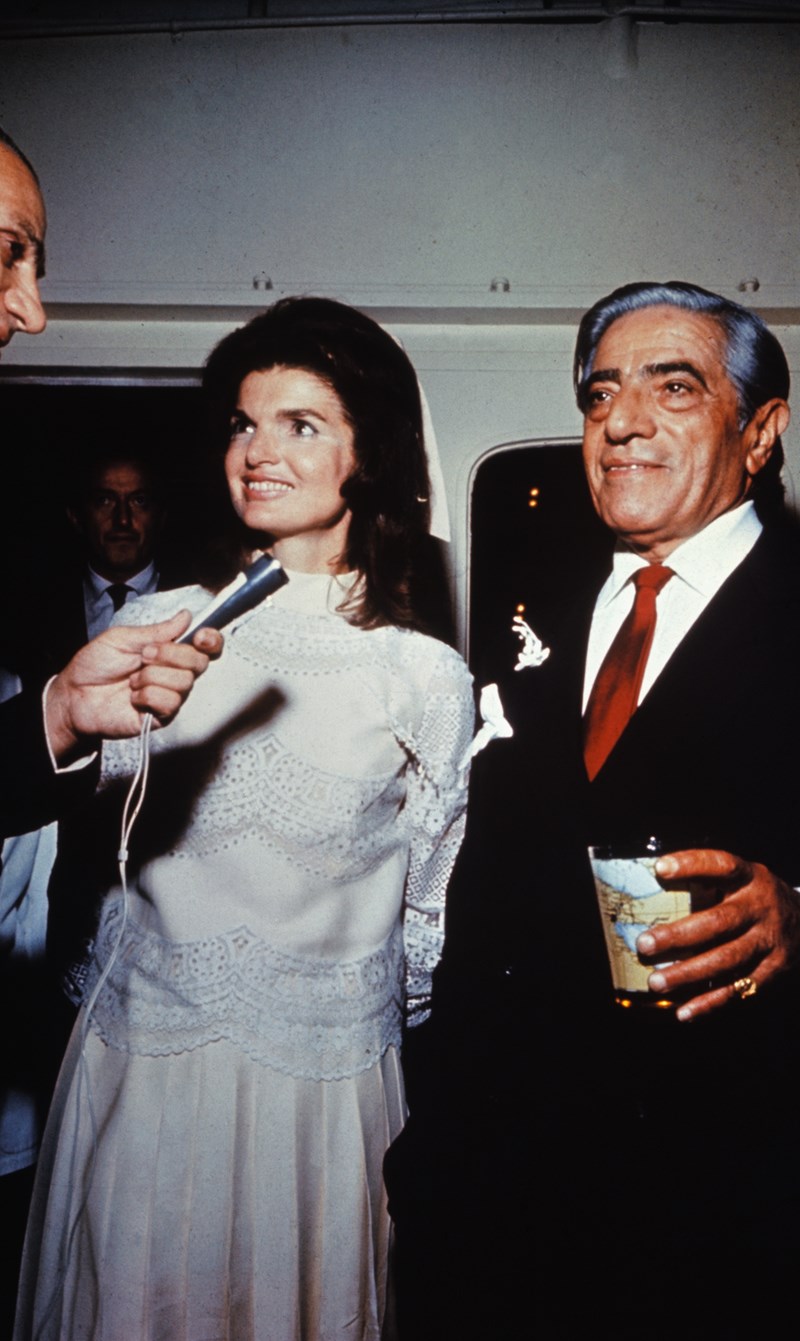 Aristotle Onassis married Jackie Kennedy on his private island. Onassis is shown with a drink in his hand and Jackie is smiling and talking into a microphone and wearing her white wedding dress.